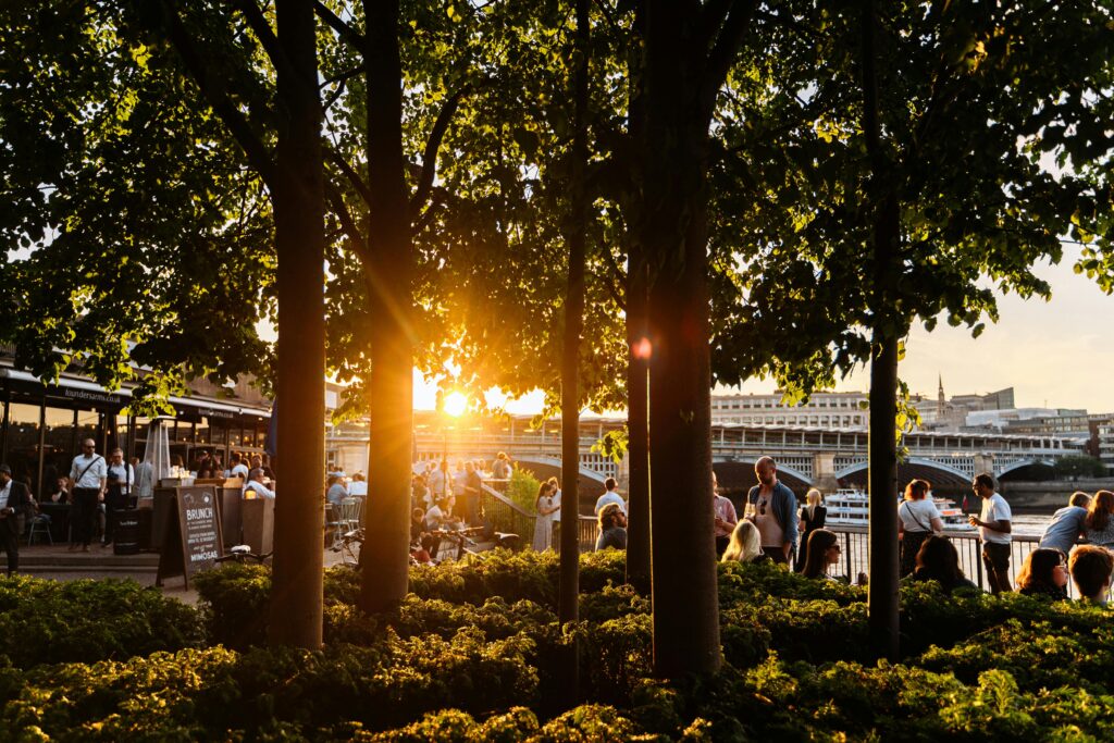 People enjoy a summer evening by the Thames River in London, with lush greenery and sunset views.